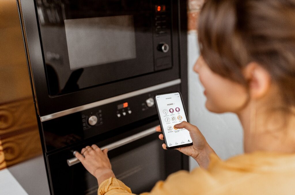 Frau bedient Handy mit Smart-Home-Steuerung, eine Hand am Ofen