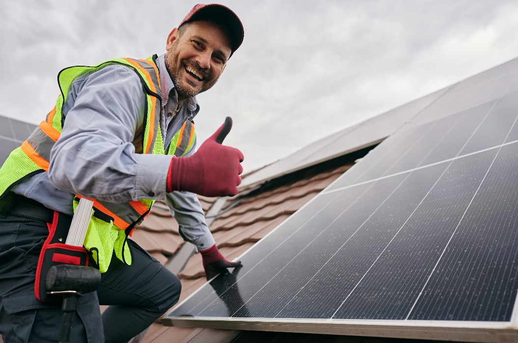 Ein zufriedener Arbeiter, der beim Anbringen von Solarmodulen auf dem Dach den Daumen hochhält und in die Kamera schaut